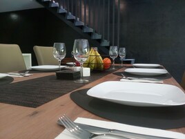 Uma mesa de jantar posta com pratos, talheres e copos de vinho, com uma cesta de frutas e uma escadaria ao fundo.