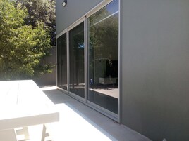 Um edifício moderno com grandes portas de correr de vidro, um pátio de concreto e uma árvore ao fundo.