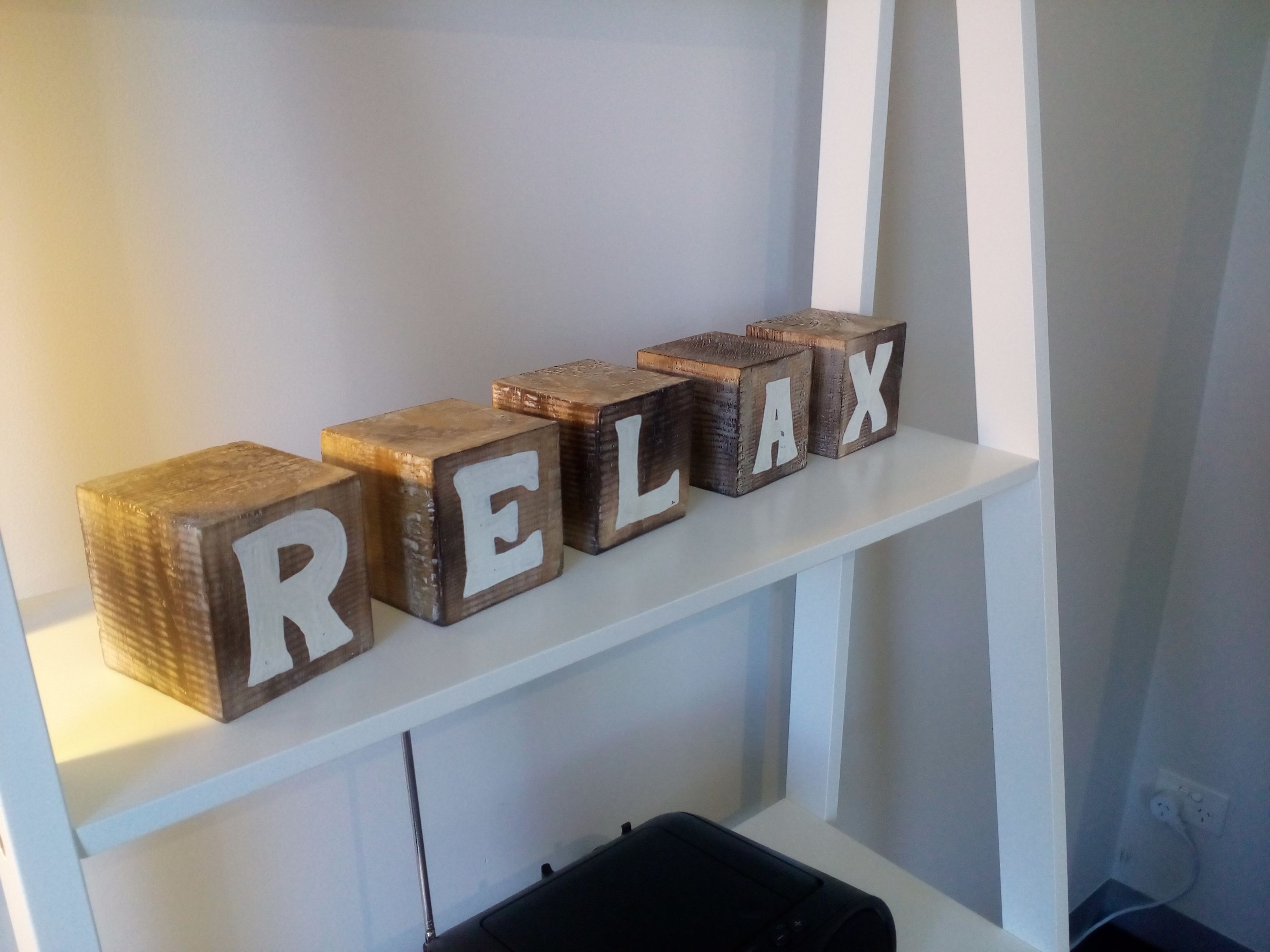 Träklossar med bokstäver som tillsammans bildar ordet ”RELAX”, placerade på en vit hylla.