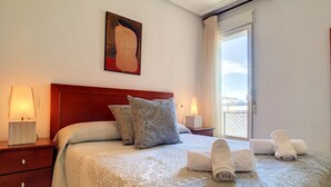 A neatly made bed with a patterned bedspread, two pillows, and rolled towels on the bed. There is a nightstand with a lamp and a small table with another lamp. A painting hangs on the wall above the bed. - Penthouse, private roof terrace, balcony, Free WiFi Internet, pool (Mar de Cristal)