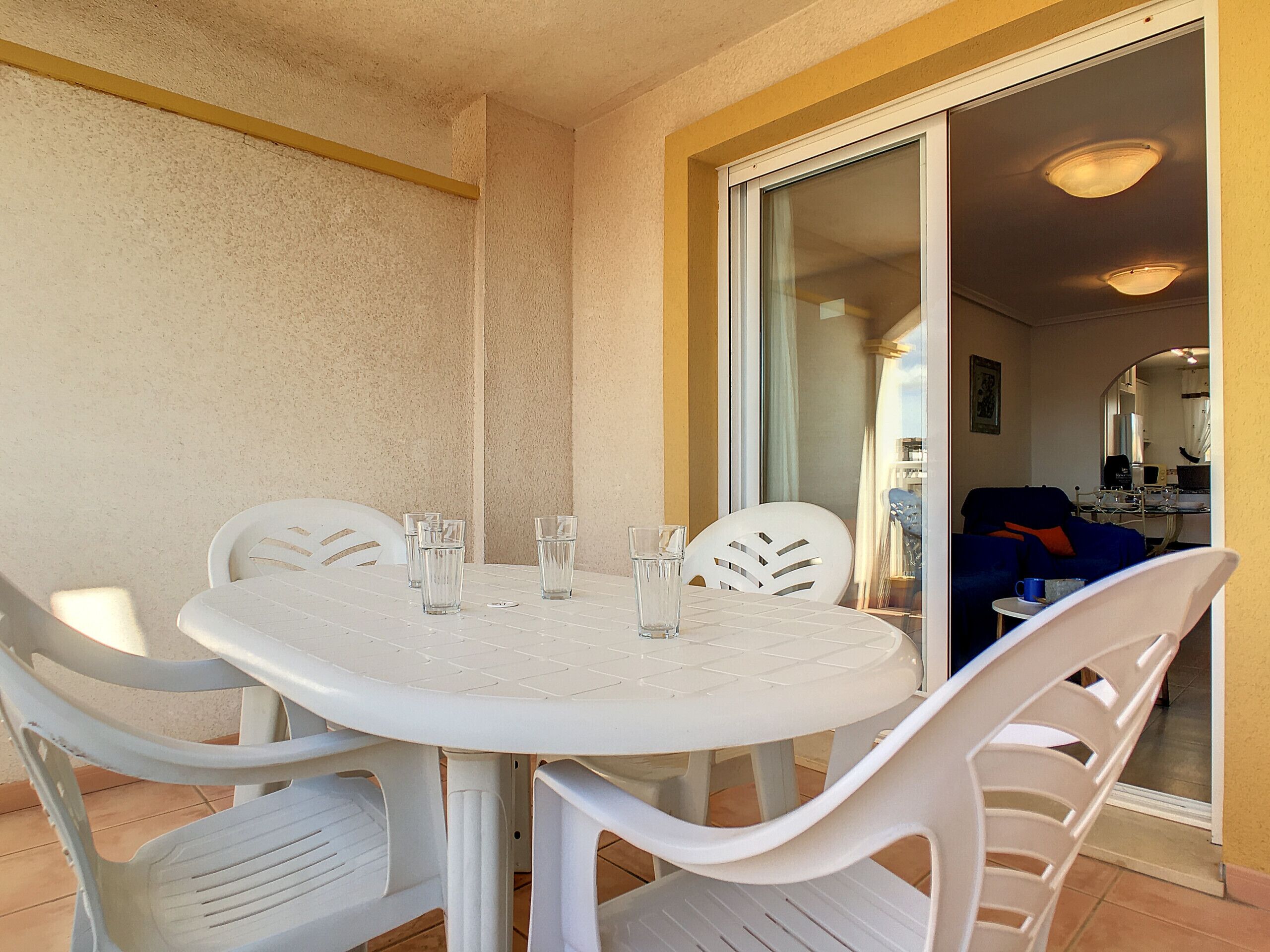 A small, private balcony with a white table and chairs, a sliding glass door leading to a bedroom, and a view of the sky.
