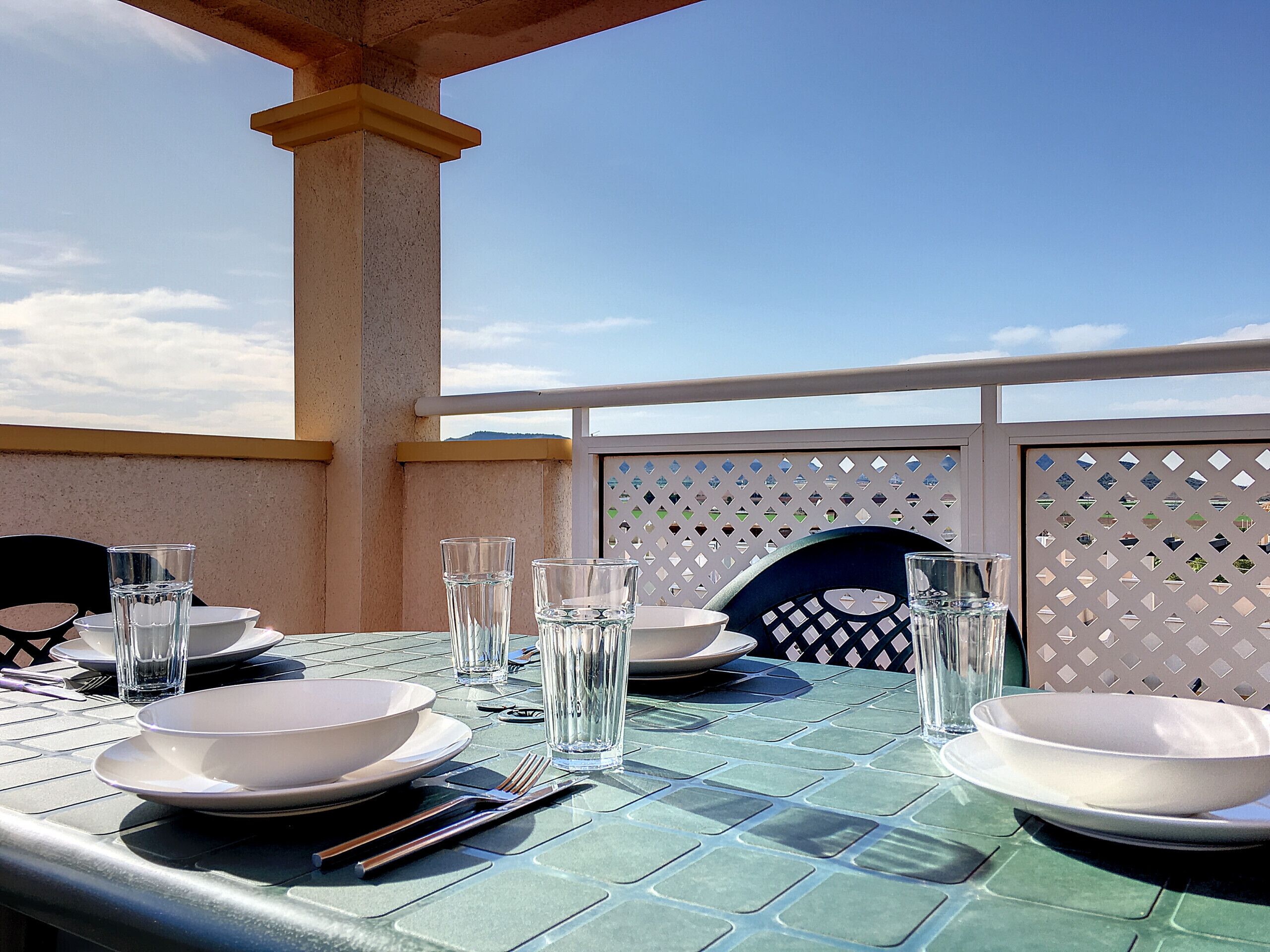 Mesa lista para comer con vasos, platos y cubiertos transparentes, bajo un balcón con vista al cielo.
