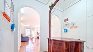 A hallway with a wooden cabinet, a sofa, and a dining table. There are framed pictures on the walls and a door with a mirror.