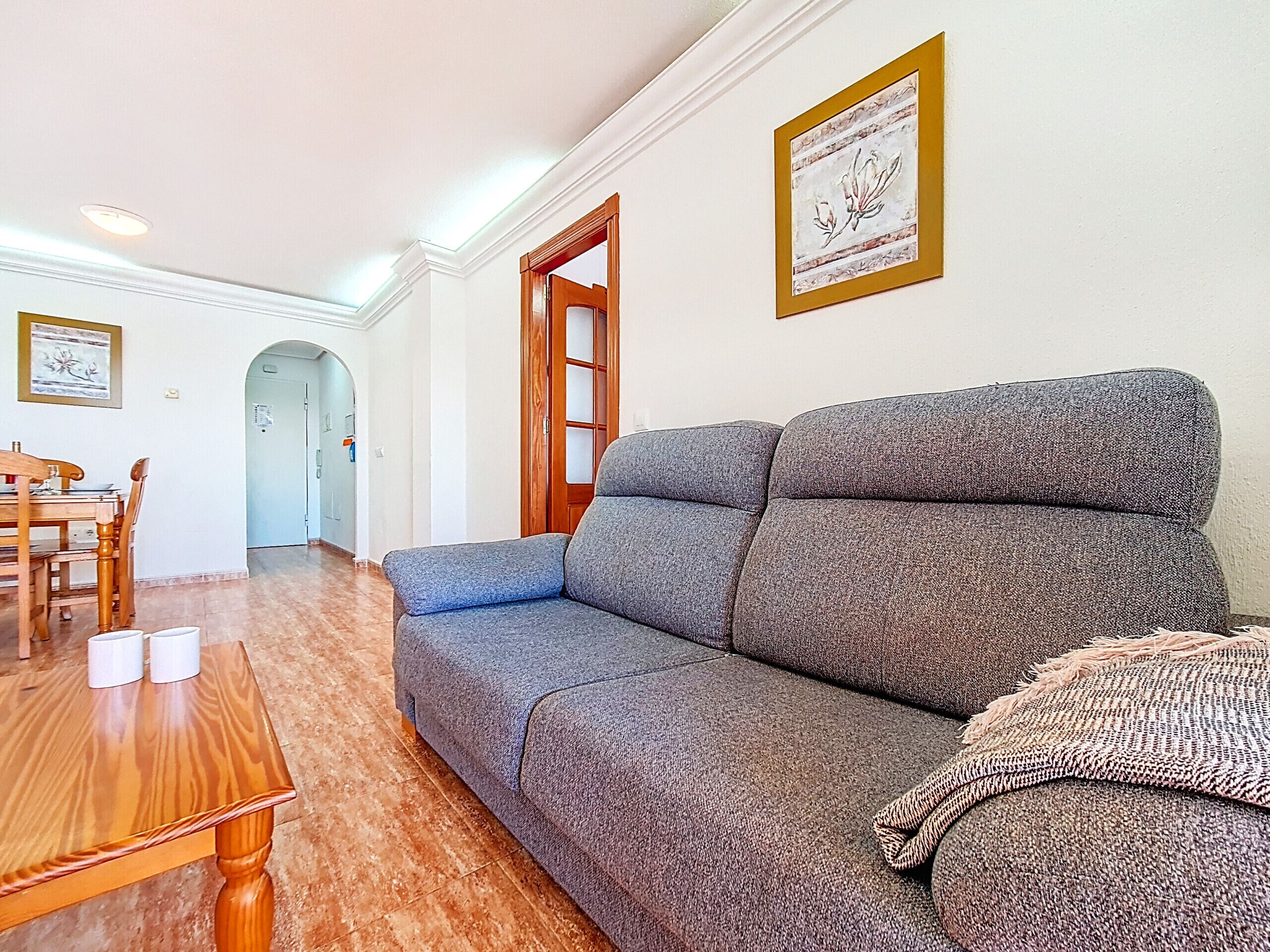 A living room with a grey sofa, wooden dining table, and chairs. There are framed artworks on the walls and a wooden door in the background.