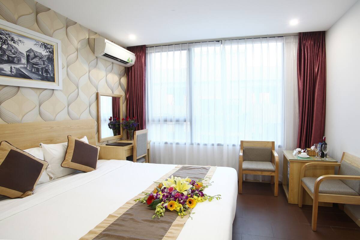 Ein Hotelzimmer mit einem großen Bett, einem Schreibtisch und einem Sessel. Es gibt ein Fenster mit Vorhängen und ein Bild an der Wand.