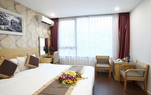 A hotel room with a large bed, a desk, and a chair. There is a window with curtains and a painting on the wall.
