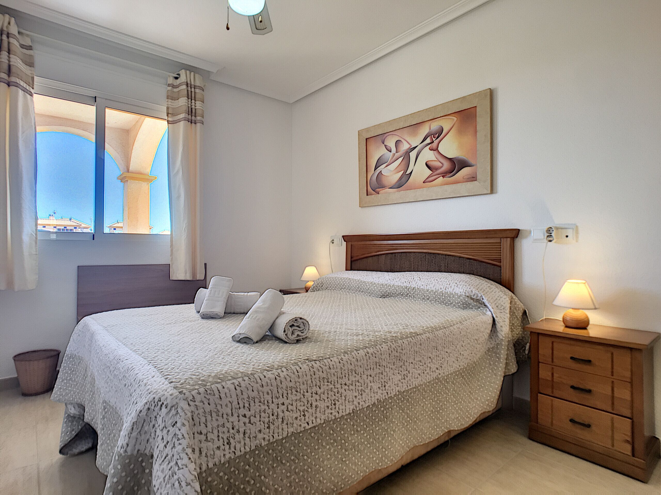 A neatly made bed with rolled towels, a wooden nightstand with a lamp, a painting on the wall, and a window with curtains.