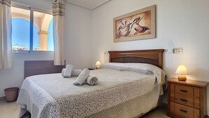 A neatly made bed with rolled towels, a wooden nightstand with a lamp, a painting on the wall, and a window with curtains.