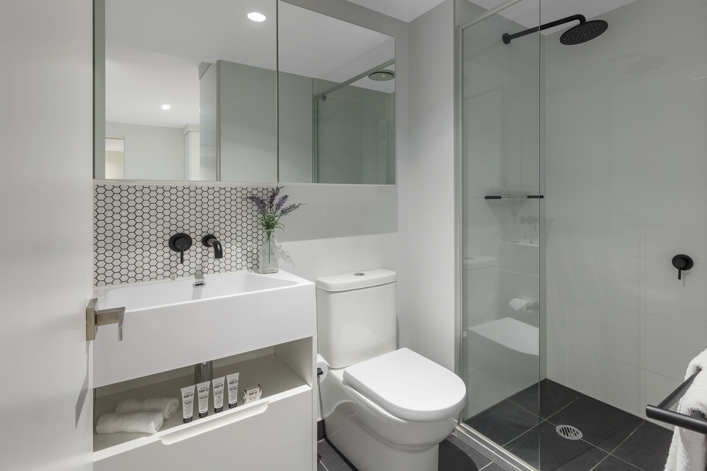 A modern bathroom with a glass shower enclosure, a white toilet, and a sink with a hexagonal tile backsplash.