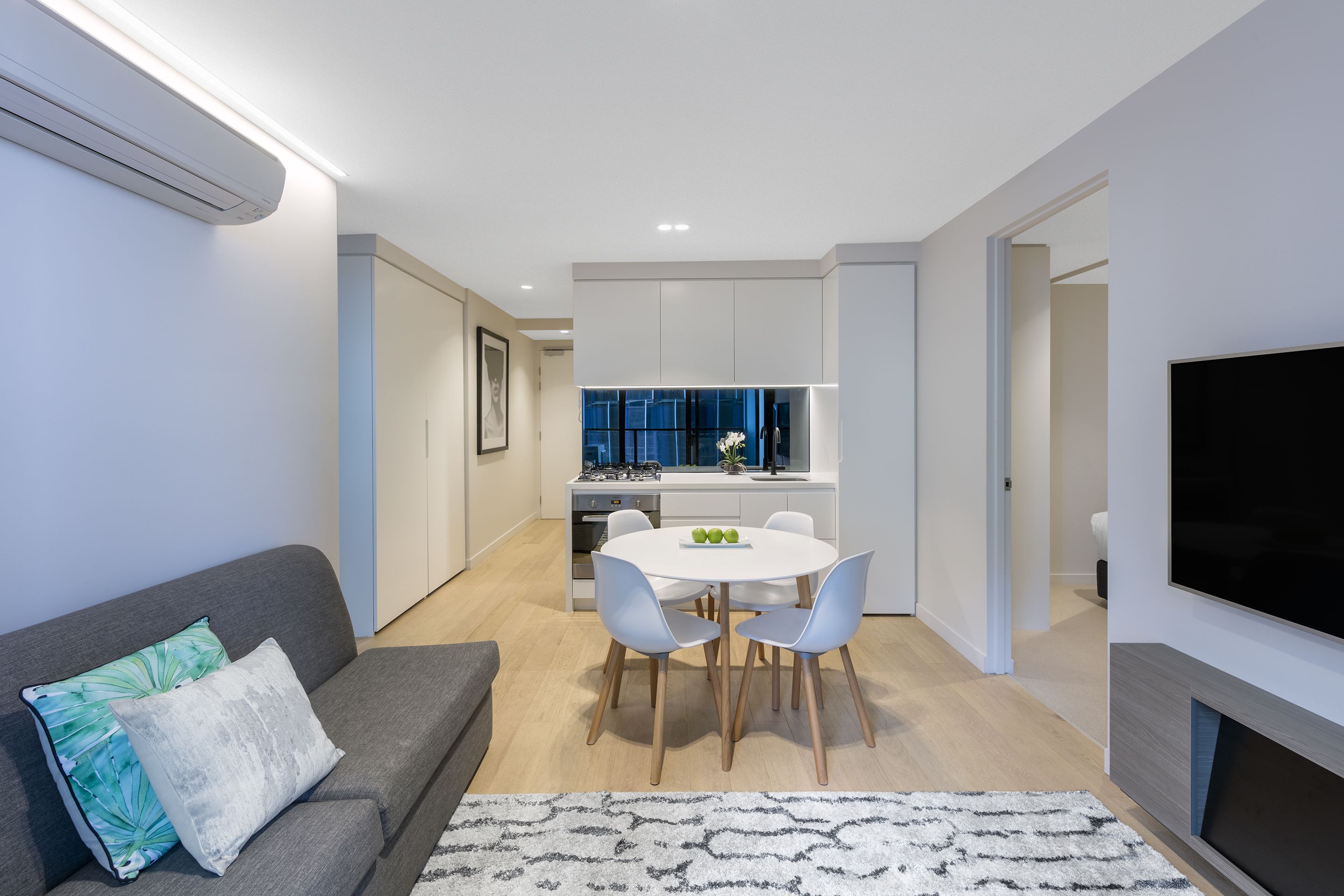 A modern living room with a grey sofa, a white dining table with four chairs, and a flat-screen TV mounted on the wall.