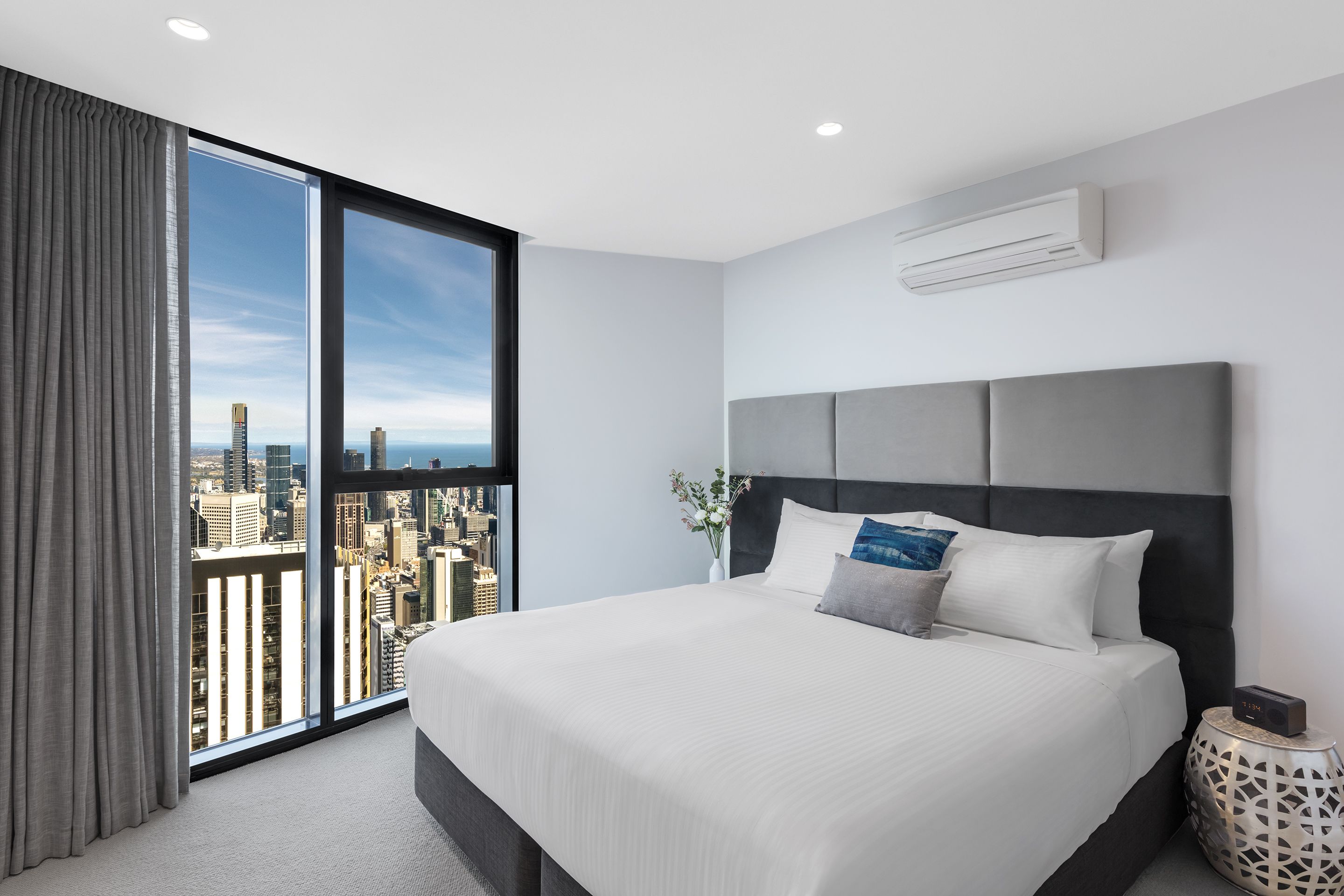 Una habitación de hotel moderna con una cama grande, vistas a la ciudad y una mesita auxiliar con un reloj.