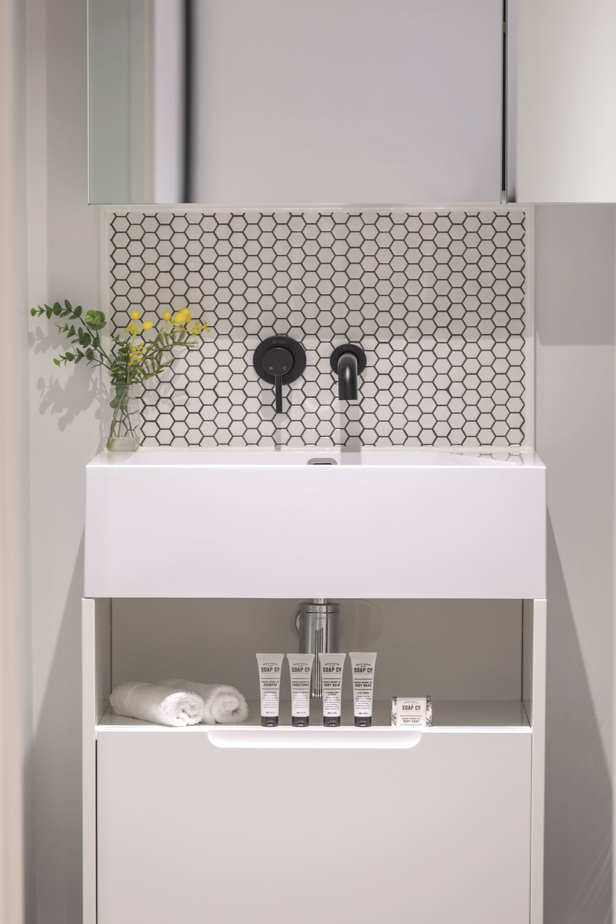 Ein modernes Badezimmer mit einem weißen Waschbecken, einer sechseckig gemusterten Spritzschutzleiste und einem Spiegel darüber. Auf dem Ablagebrett des Waschbeckens stehen Toilettenartikel.