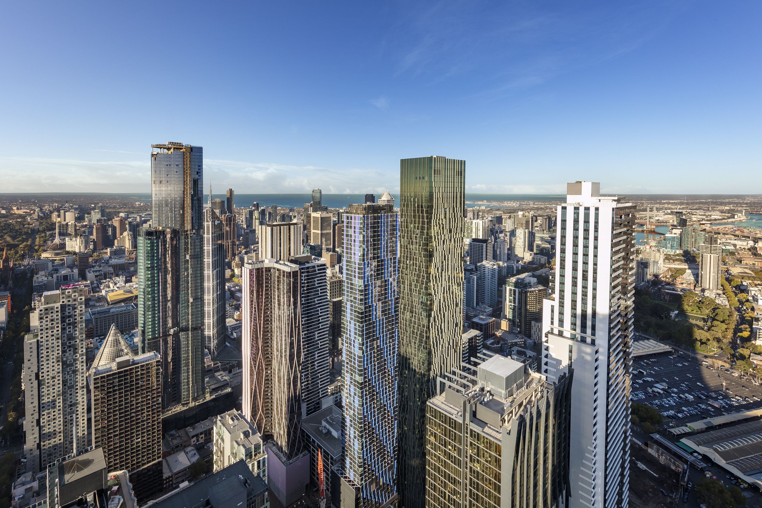 A high-rise urban skyline with modern skyscrapers, a clear blue sky, and a distant view of a body of water.