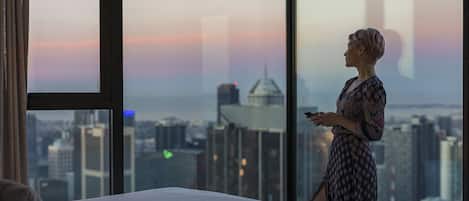 Une personne se tient près d’une grande fenêtre donnant sur une vue panoramique de la ville au crépuscule.