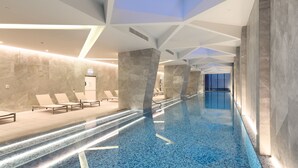 A modern indoor swimming pool with geometric ceiling design, lounge chairs, and a skylight.