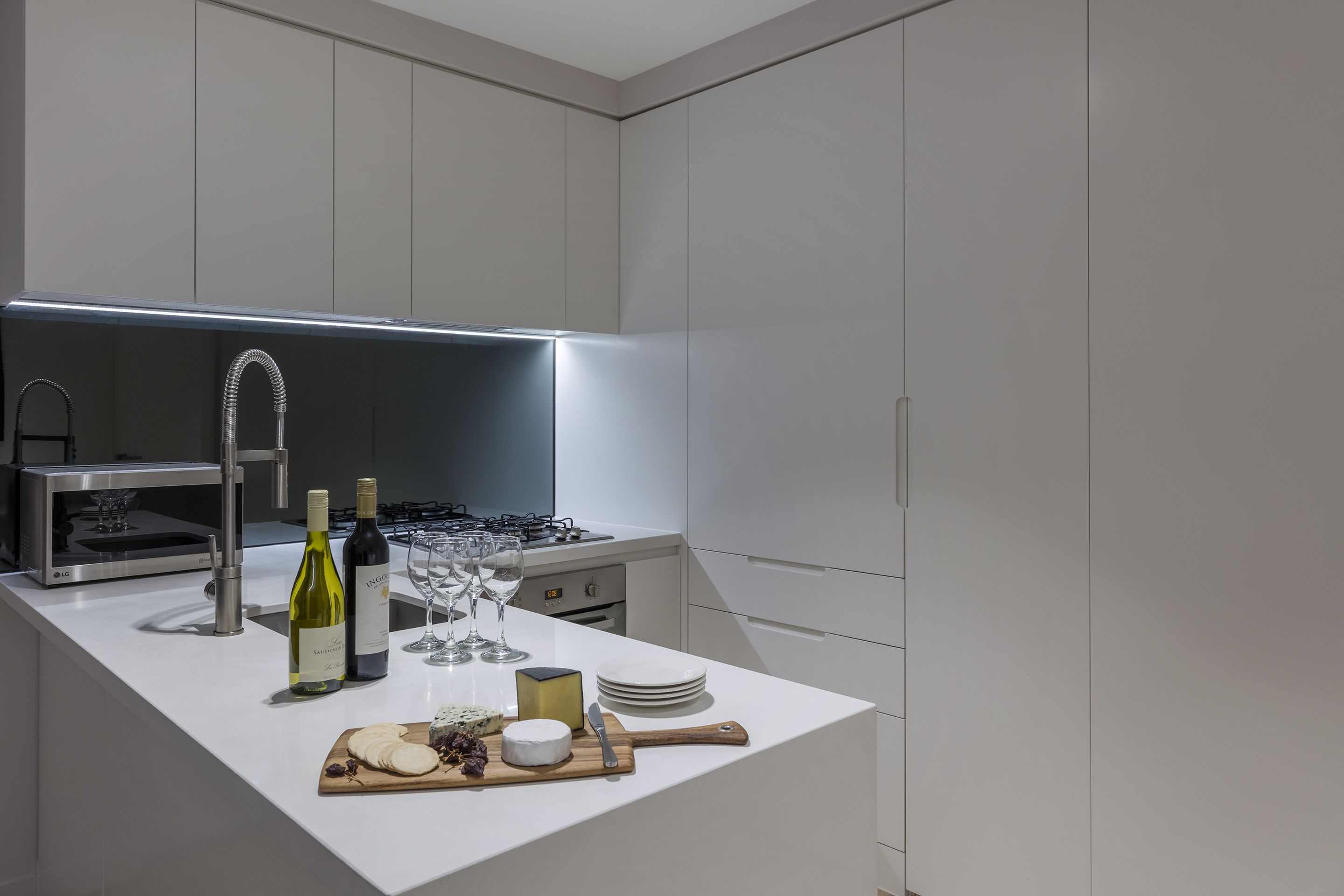 A modern kitchen with white cabinetry, a stainless steel range hood, and a countertop with wine bottles, glasses, and cheese.