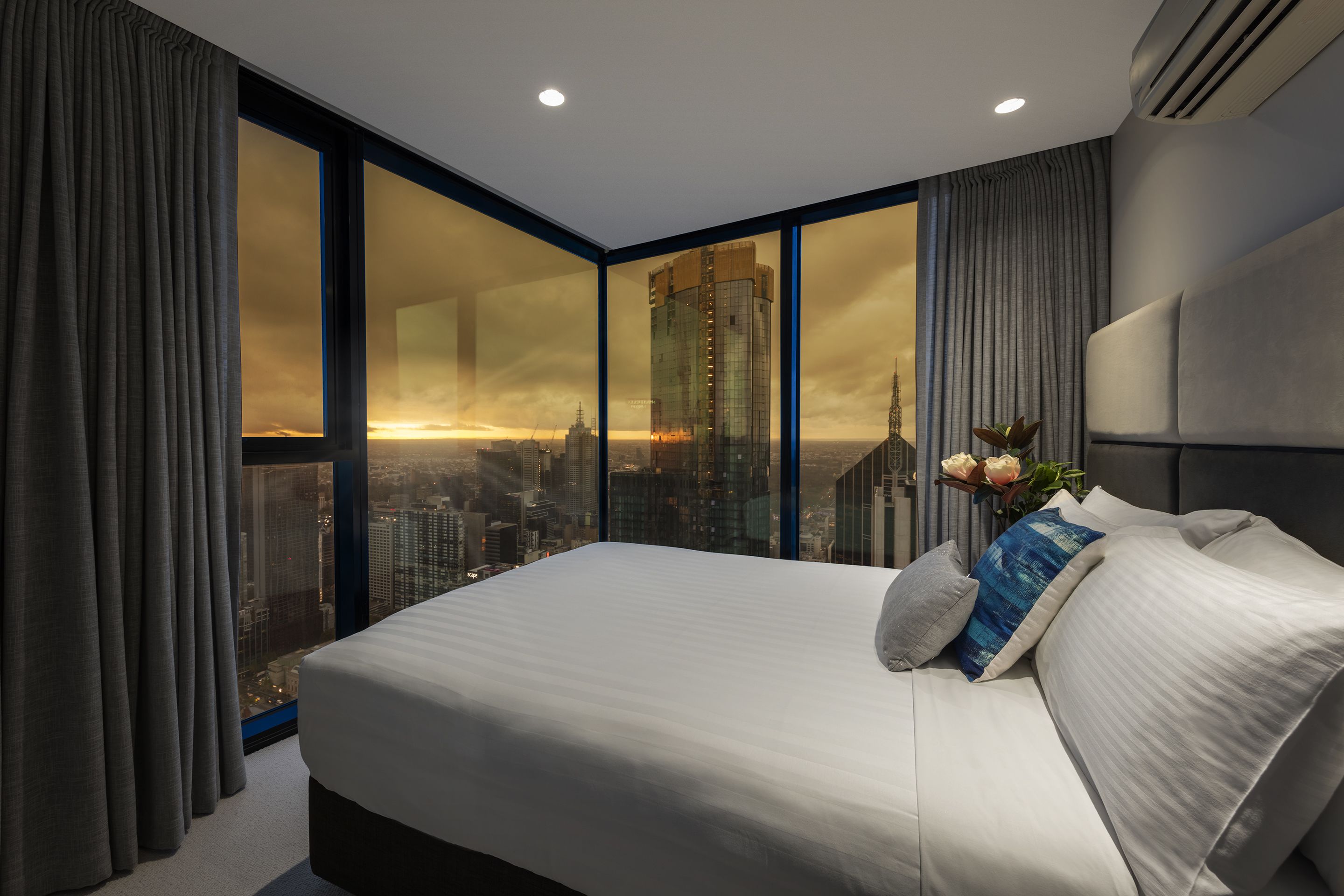A modern hotel room with a large bed, a view of a cityscape through floor-to-ceiling windows, and a city skyline at sunset.