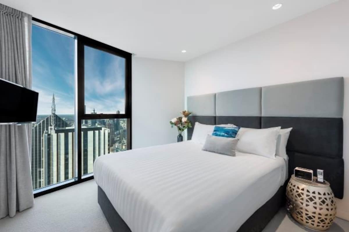 A modern hotel room with a large bed, a flat-screen TV, and a city view.