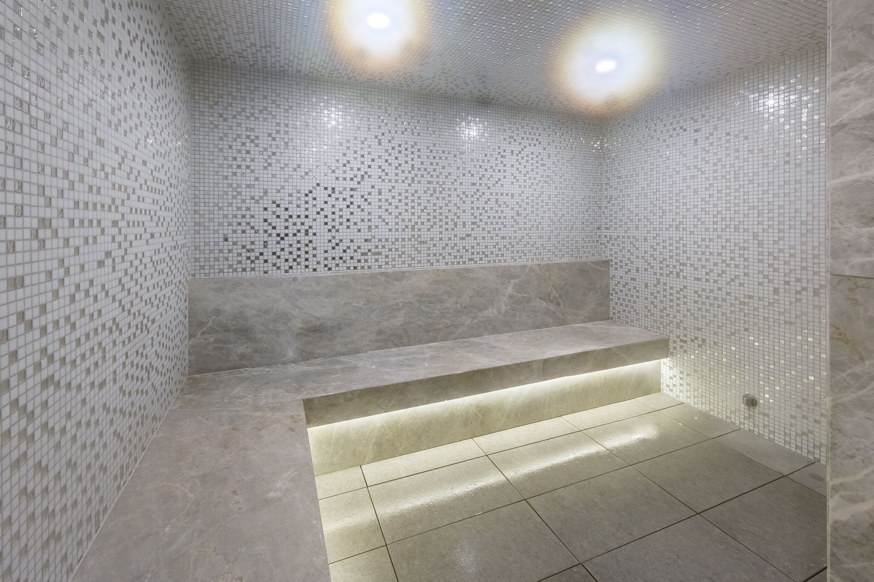 A modern bathroom with mosaic tile walls, a marble bench, and recessed lighting.