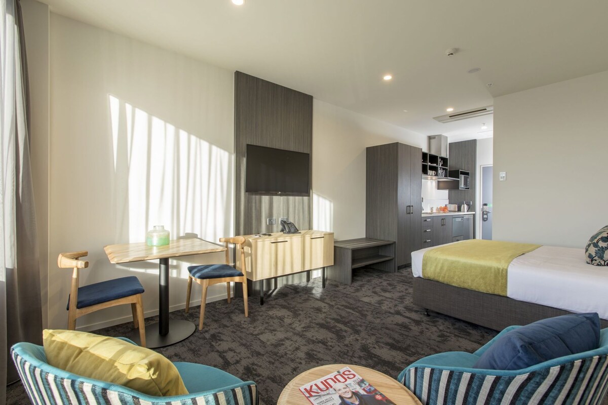 A hotel room with a bed, desk, chair, kitchenette, and a small seating area.
