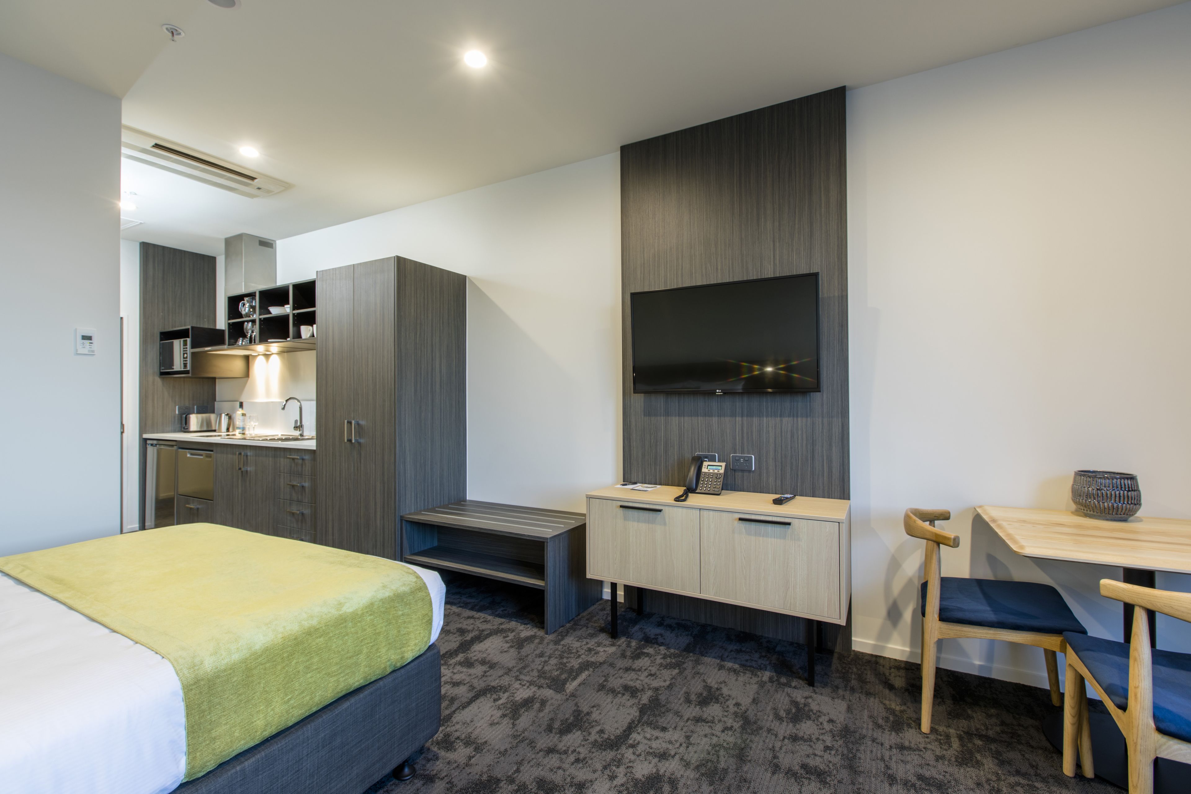 Una habitación de hotel moderna con cama, escritorio, silla, una kitchenette pequeña y televisor montado en la pared.