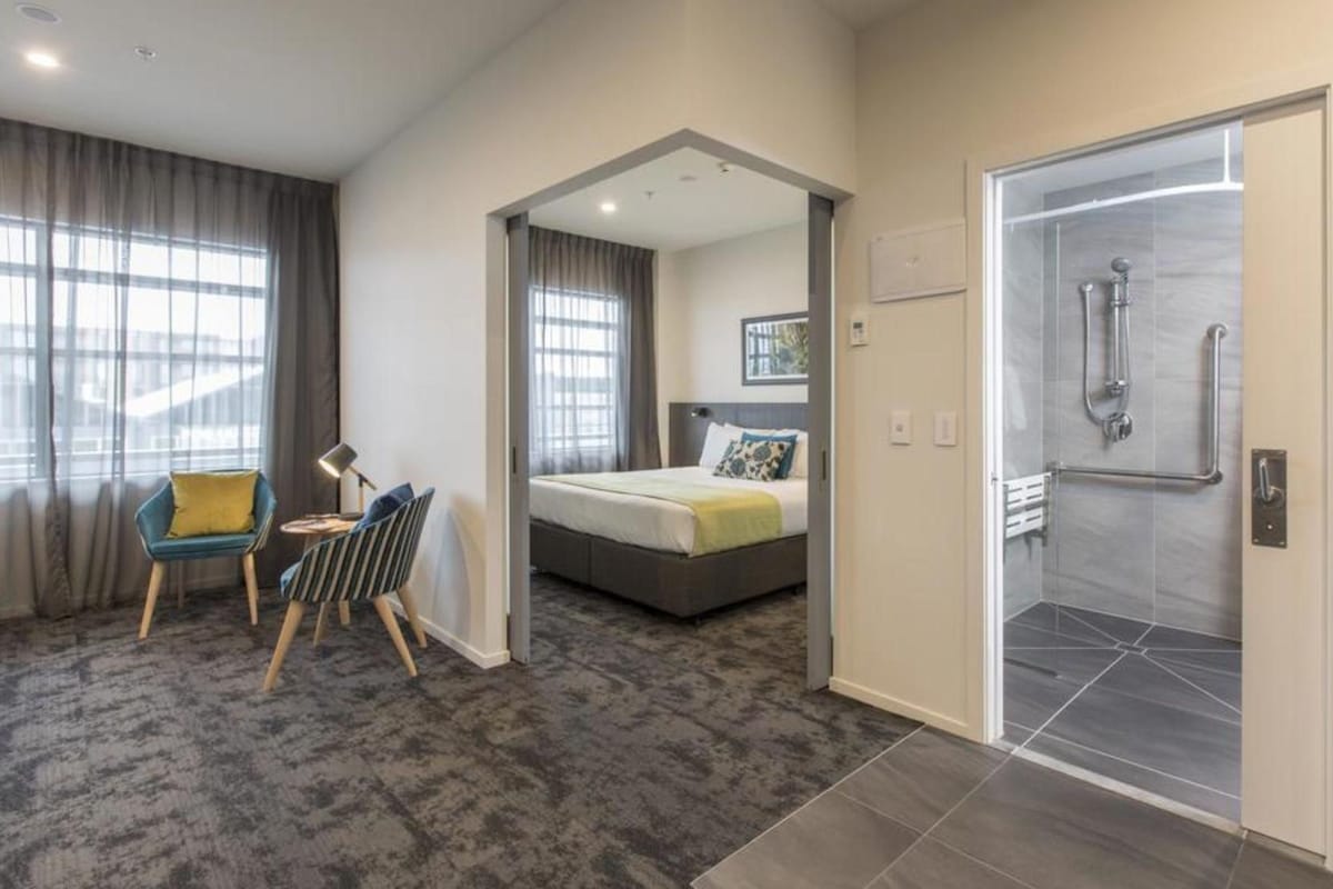 A modern hotel room with a bed, a desk, a chair, and a shower area.