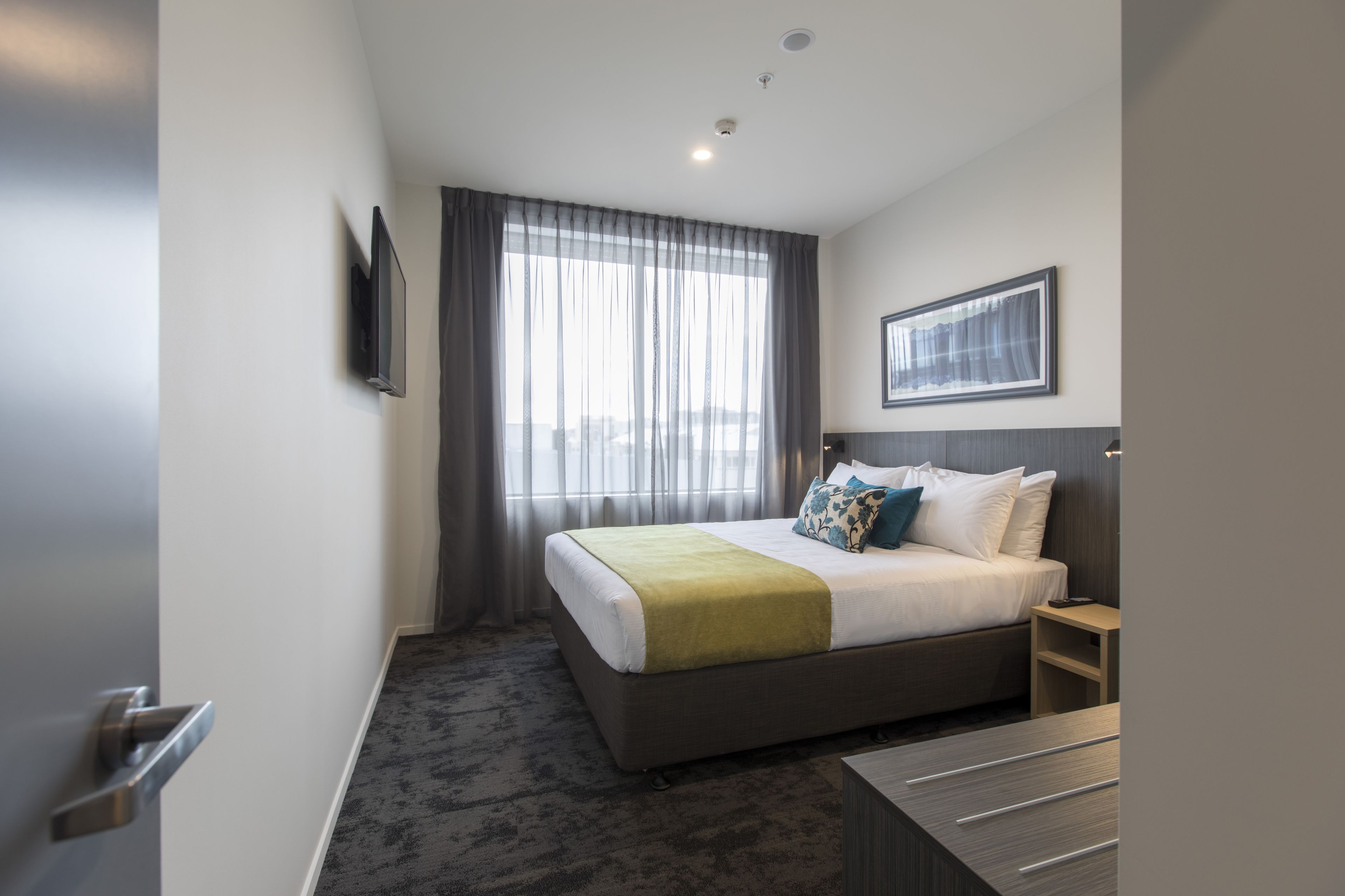 Habitación de hotel con cama, mesita de noche, televisor montado en la pared y ventana con cortinas.