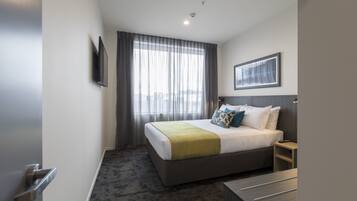 A hotel room with a bed, a bedside table, a TV mounted on the wall, and a window with curtains.