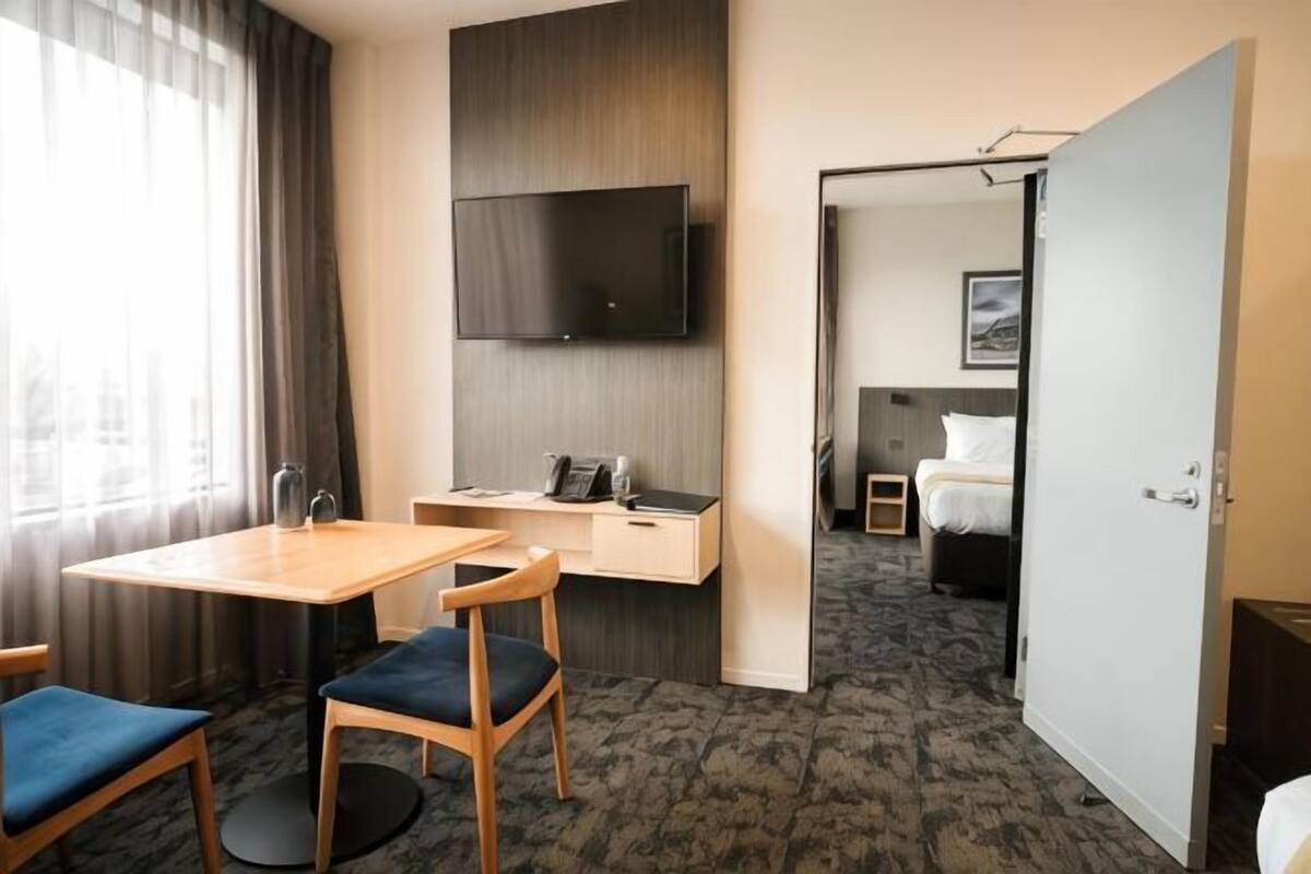 A hotel room with a desk, chairs, a flat-screen TV, a bed, and a door leading to another room.