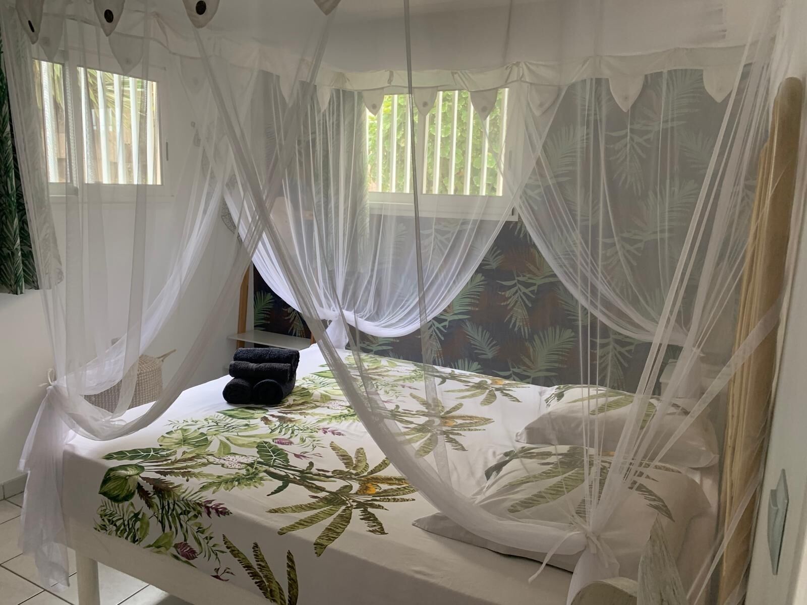 A room with a bed under a canopy, a small table with towels, and a window with curtains.