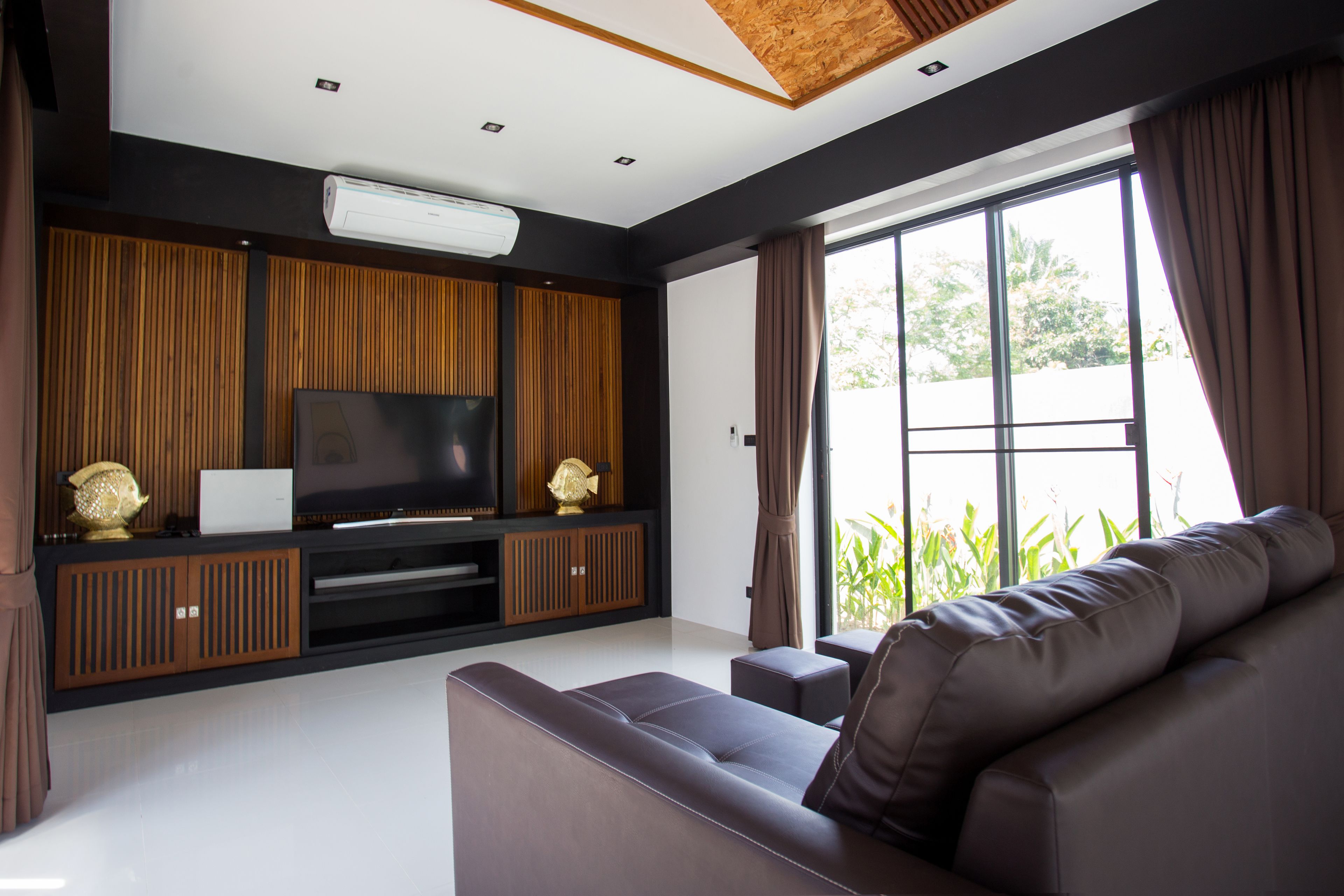 A modern living room with a large flat-screen TV, wooden paneling, a sofa, and a large window with curtains.