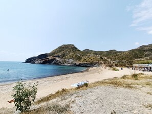 Une plage avec des collines rocheuses en arrière-plan, une rive sablonneuse et quelques personnes ainsi que des parasols.