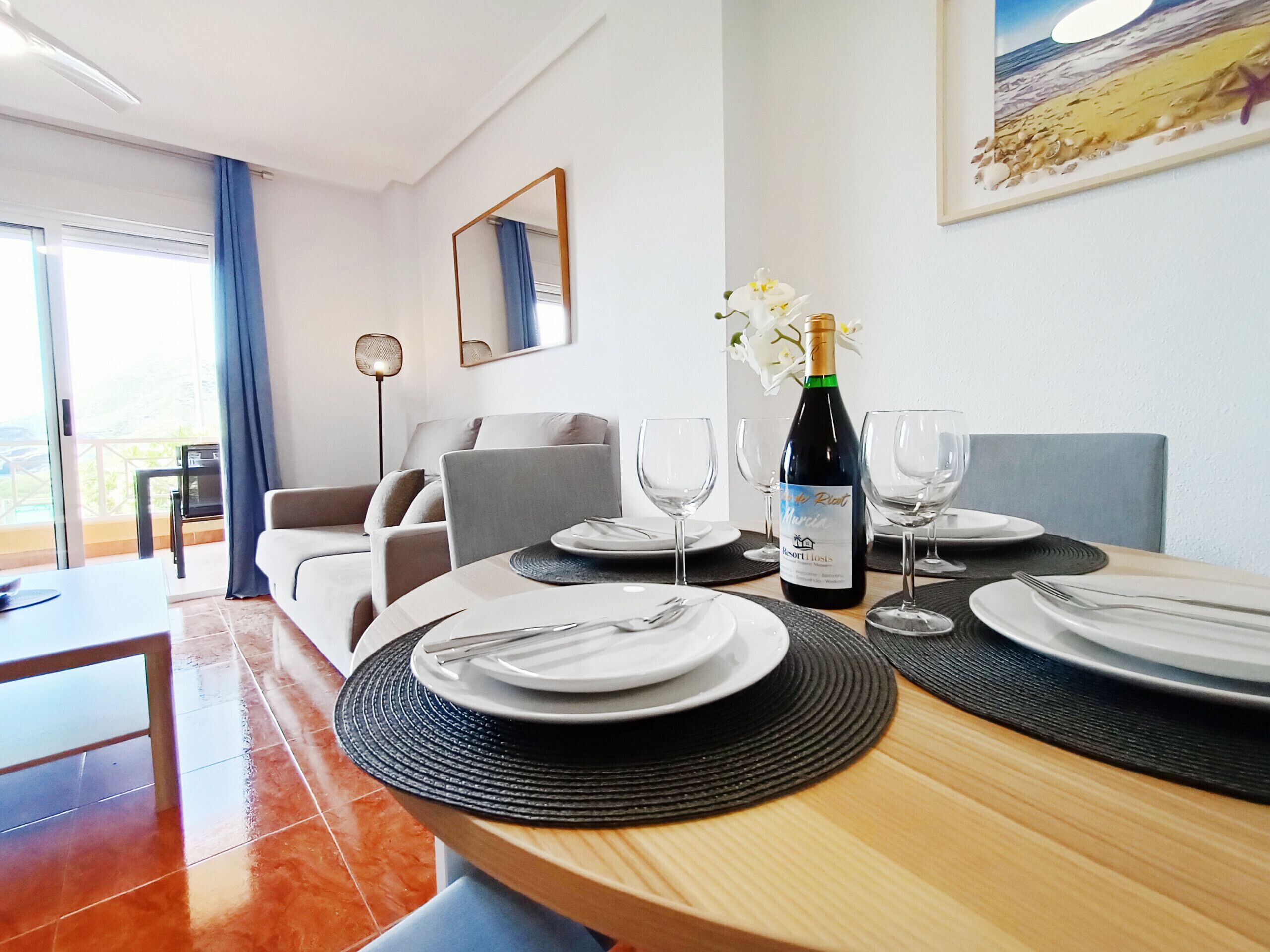 Un comedor con una mesa de madera dispuesta para cuatro personas, una botella de vino y un cuadro en la pared.