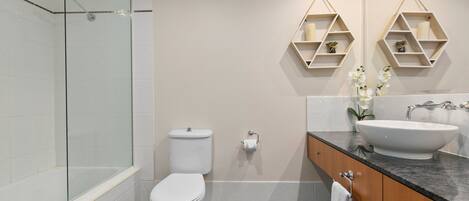 A bathroom with a glass-enclosed shower, a toilet, a bathtub, and a vanity with a sink and mirror. There are two geometric shelves on the wall.