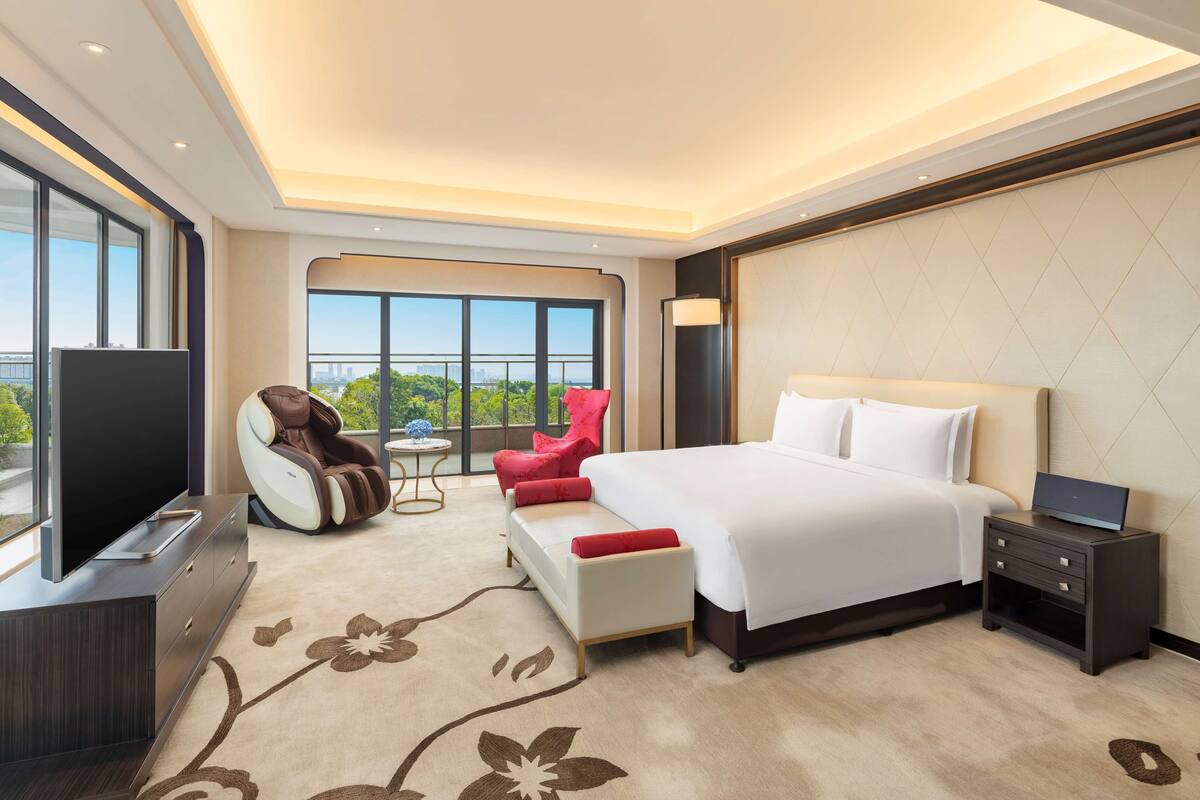 Une chambre d’hôtel moderne avec un grand lit, un téléviseur à écran plat, un fauteuil rouge et une vue sur la ville grâce à de grandes fenêtres.
