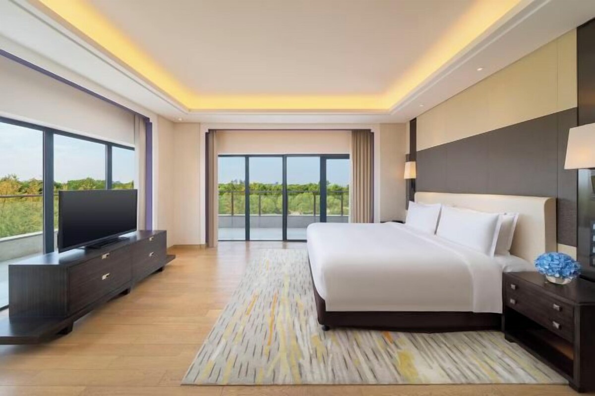 A modern bedroom with a large bed, a flat-screen TV on a dark wooden console, and a view of greenery through large windows.