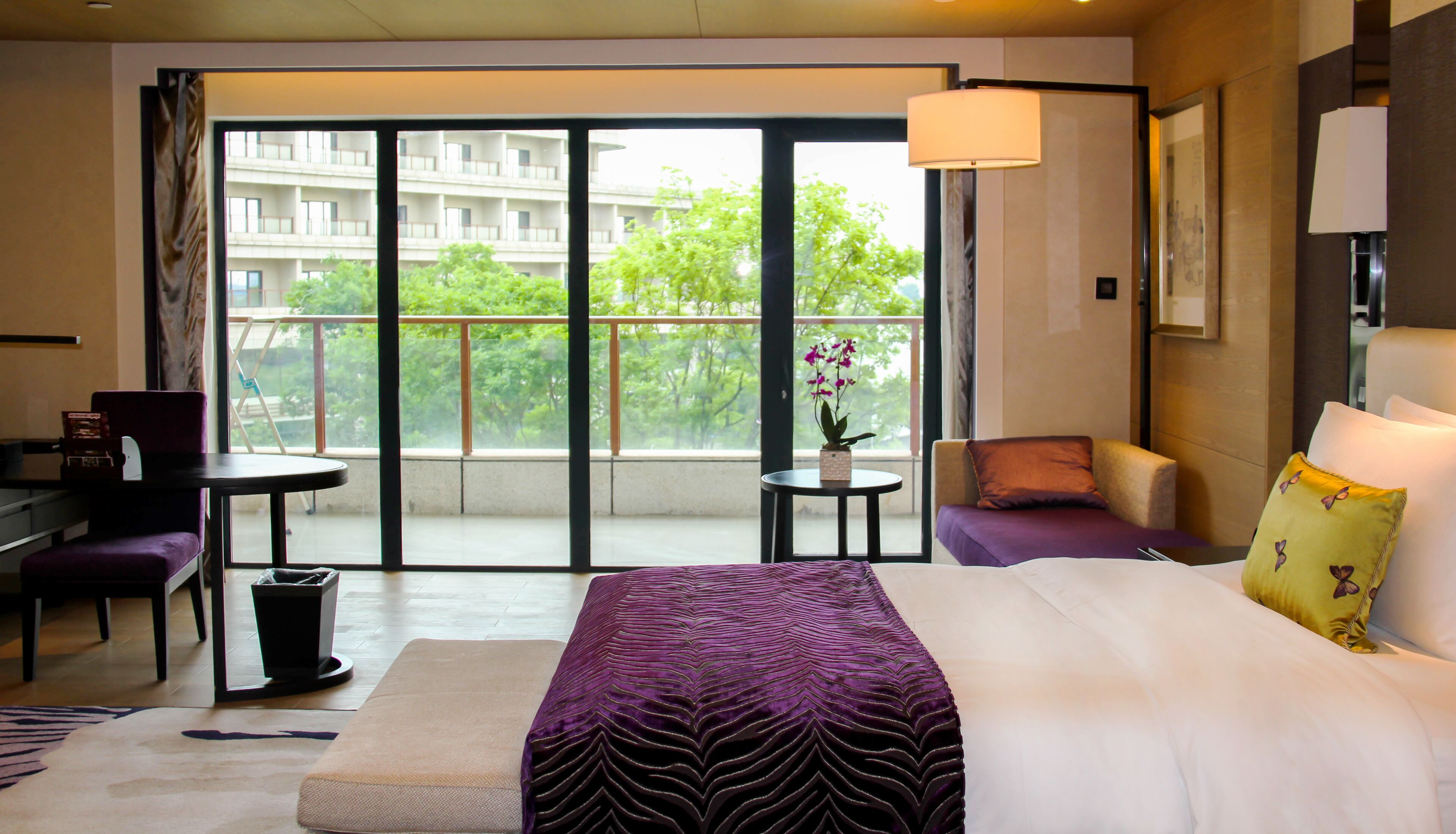 A hotel room with a large bed, a desk, a chair, a small table with a flower, and a view of a building and trees through the balcony doors.