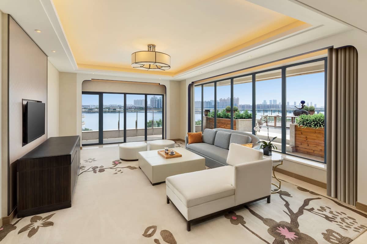 Un salon moderne avec un canapé, deux fauteuils blancs, une table basse et un téléviseur posé sur un meuble sombre. De grandes fenêtres offrent une vue sur les toits d’une ville et sur une étendue d’eau.
