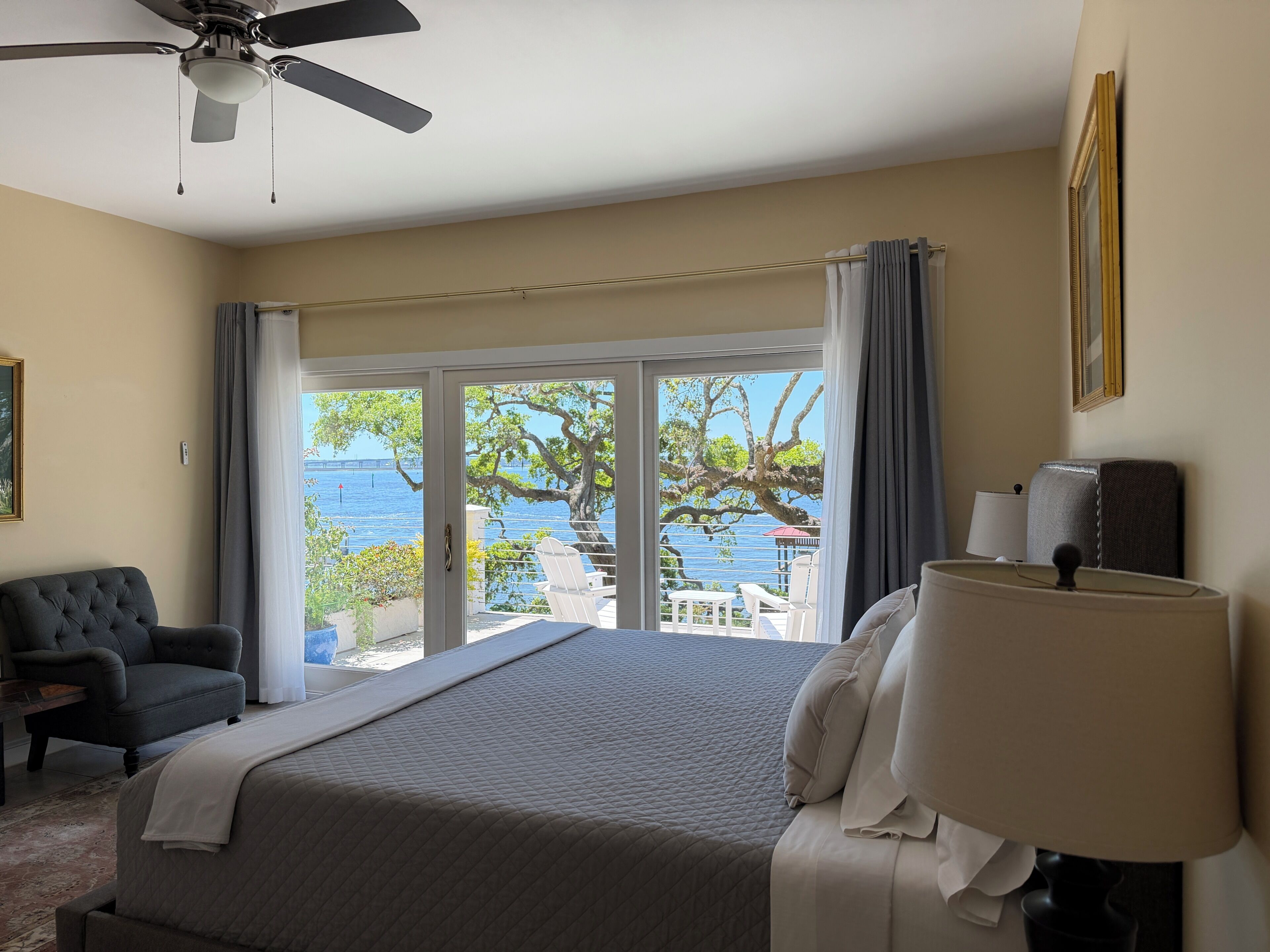 A bedroom with a bed, a chair, a lamp, a ceiling fan, and a view of the sea through a sliding door.