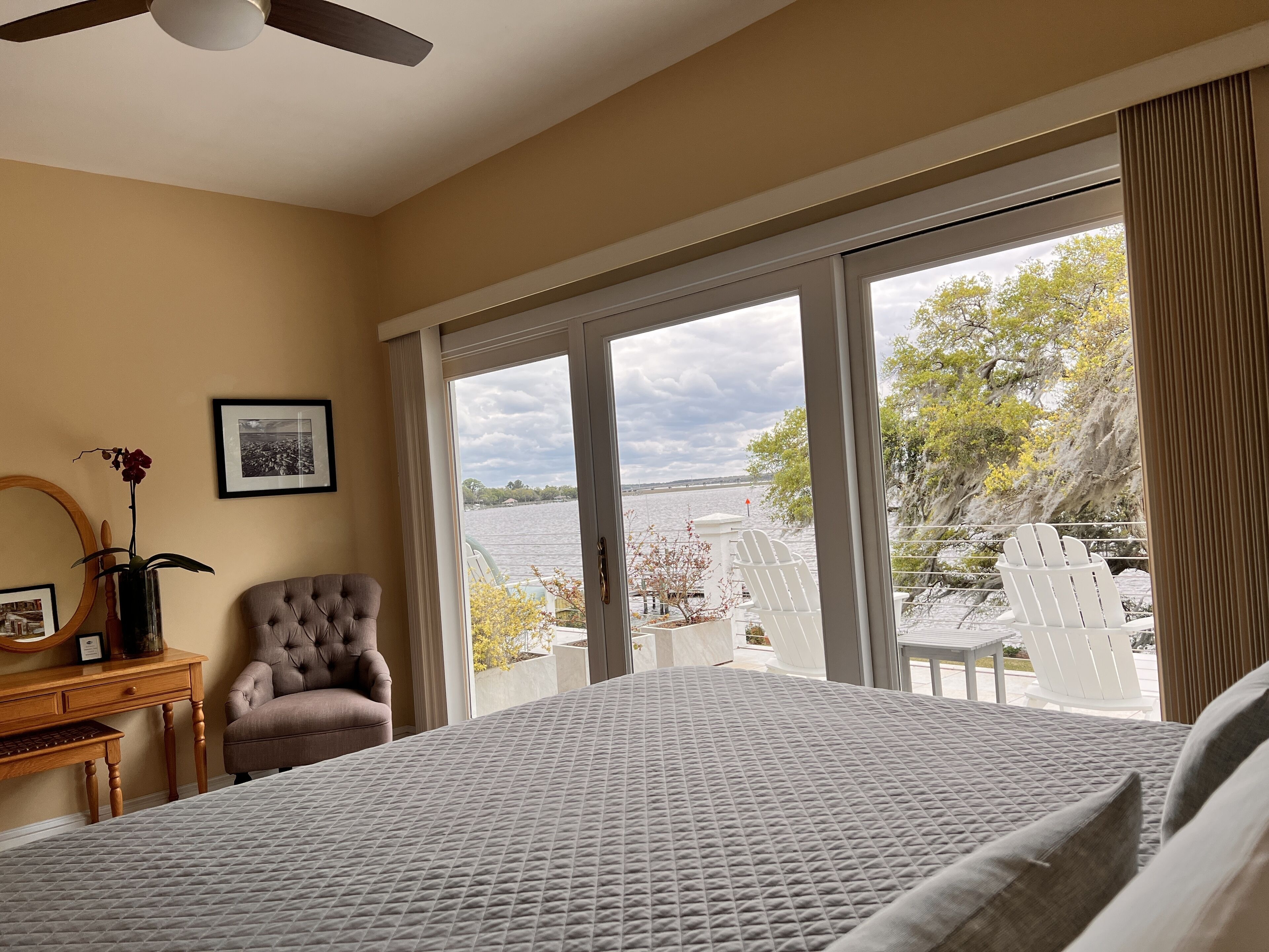 A bedroom with a large bed, a chair, a desk, and a view of a body of water through large windows.