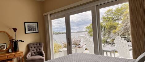 A bedroom with a large bed, a chair, a desk, and a view of a body of water through large windows.