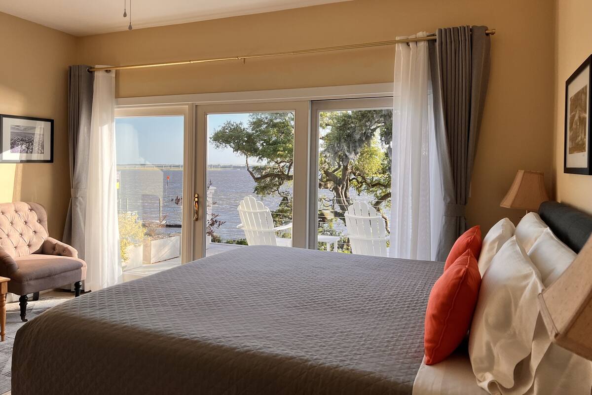 Une chambre avec un grand lit, une chaise, un ventilateur de plafond et une vue sur une étendue d’eau, visible à travers une porte-fenêtre coulissante.