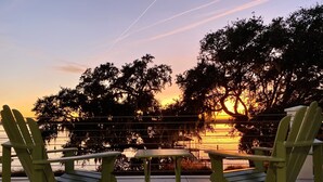 A sunset view with two Adirondack chairs and a small table on a concrete deck, overlooking a body of water and trees.