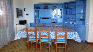 A dining area with a table and chairs, a blue cabinet with shelves, and a small television on a white cabinet. - Large panoramic apartment, beautiful sea view. (Costa Rei)