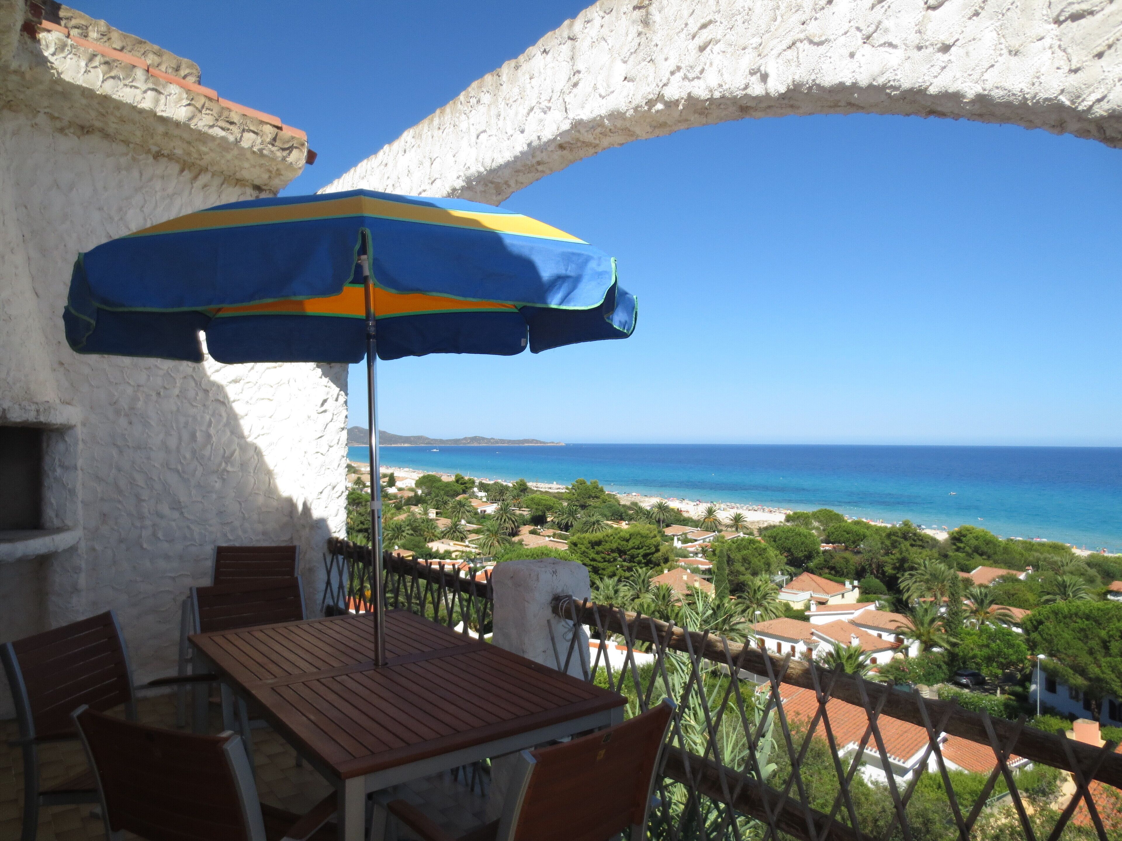 Un balcon avec une table et des chaises en bois, un parasol et une vue sur une plage et l’océan.
