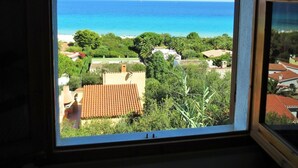 A view from a window with a curved frame, overlooking a coastal area with buildings and a clear blue sky. - Large panoramic apartment, beautiful sea view. (Costa Rei)