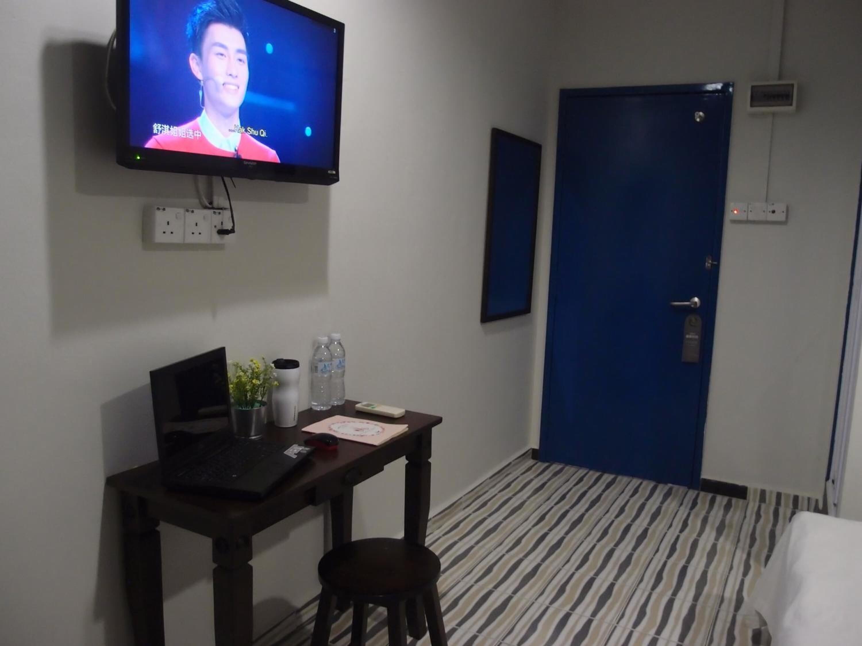 Habitación de hotel con televisión montada en la pared, un escritorio con una computadora portátil, una silla, una mesita con un florero y botellas de agua, y una puerta azul.