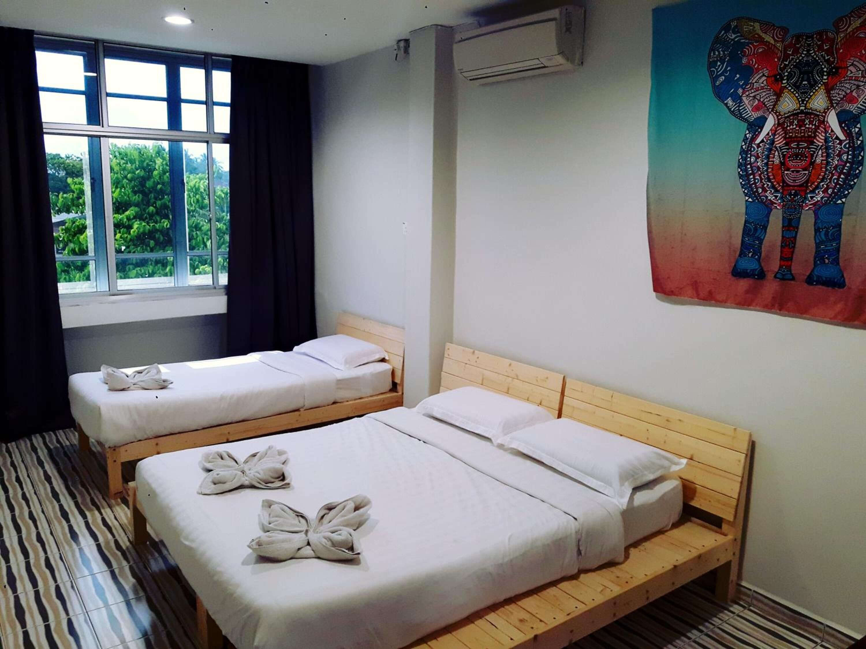 A hotel room with two single beds, a window with curtains, an air conditioning unit, and a colorful elephant painting on the wall.