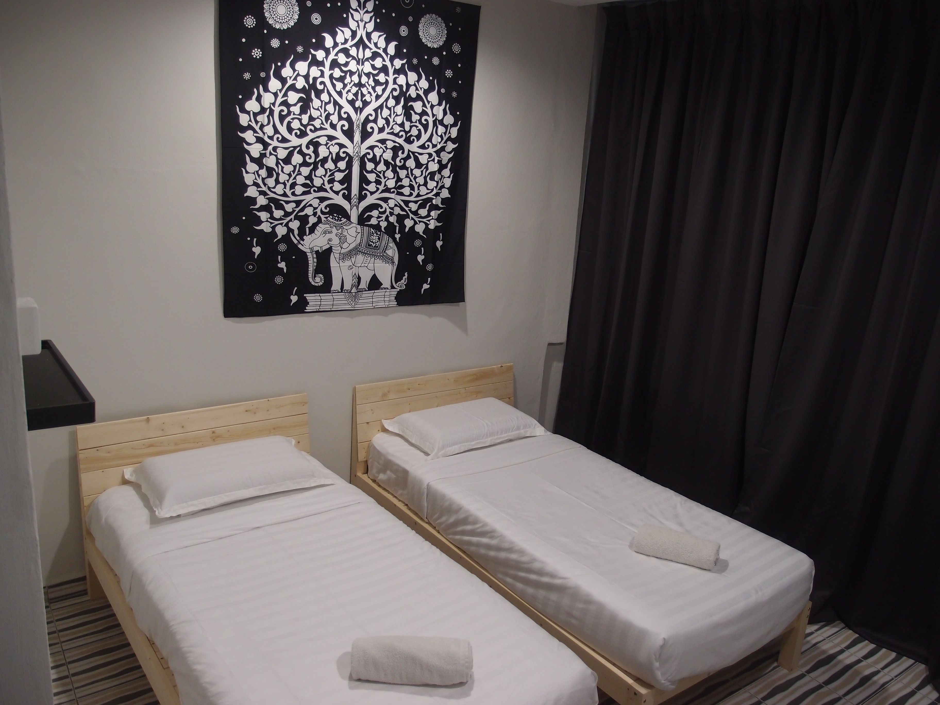 A room with two single beds, a black tapestry with a tree and elephant design, a wooden headboard, a black nightstand, and a black curtain.