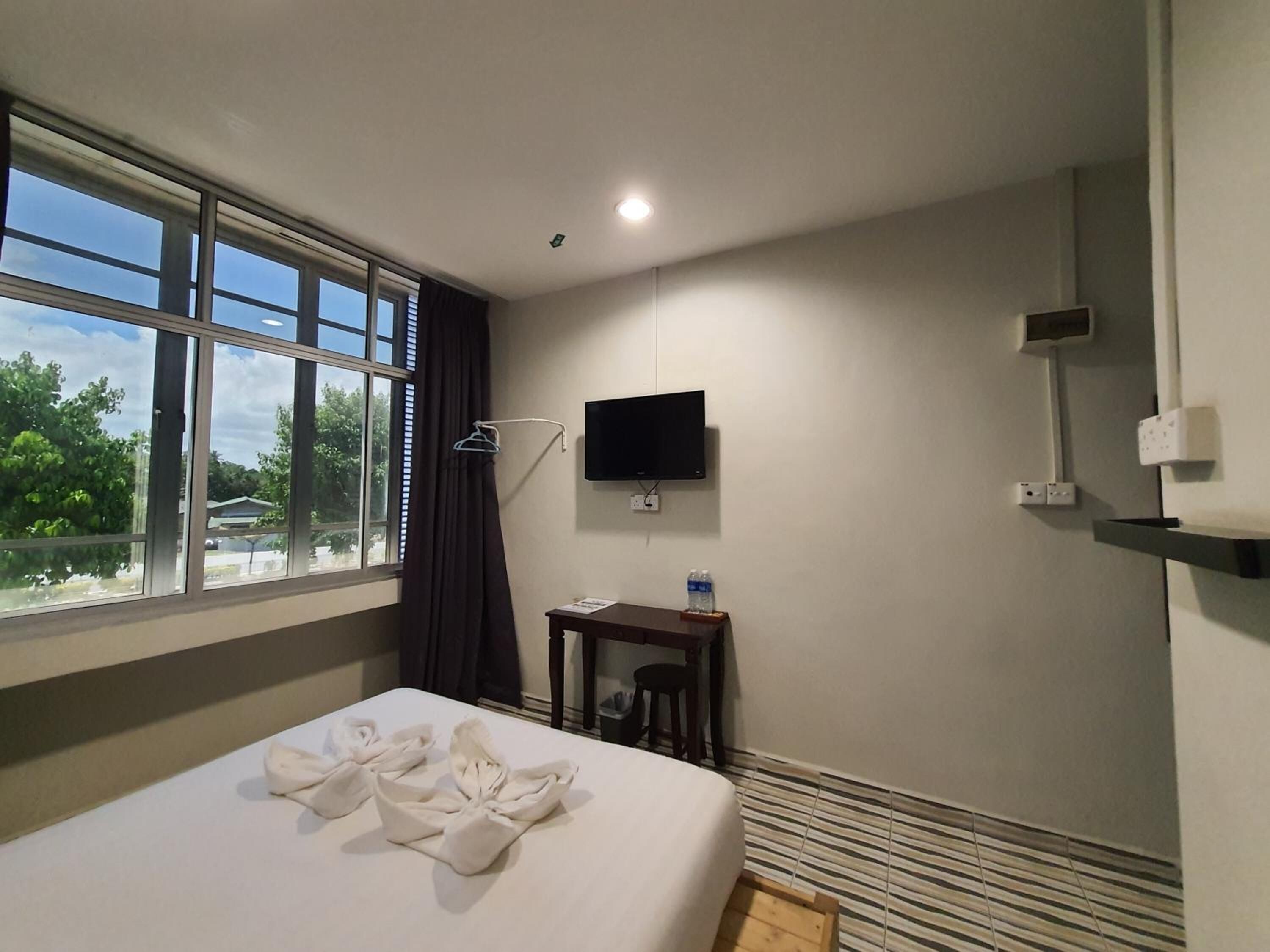 A hotel room with a bed, a TV mounted on the wall, a small table with a bottle, and a window with curtains.